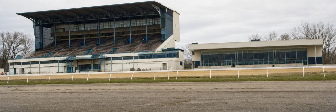 A racetrack grandstand of painted steel and concrete standing beside a lower, newer gaming building, seen across an empty asphalt apron under an overcast sky, with a white running rail between them. (AI-generated image)
